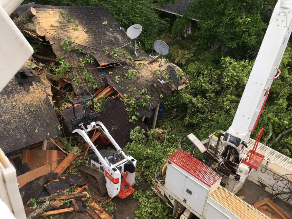 An aerial view of a damaged roof with tree debris, a Bobcat skid steer, and a crane truck performing storm cleanup by Tri-County Tree And Restoration in Jackson, MS.