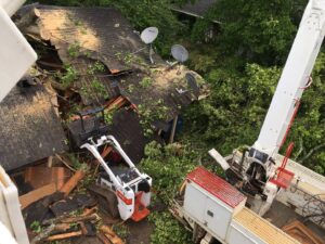 An aerial view of a damaged roof with tree debris, a Bobcat skid steer, and a crane truck performing storm cleanup by Tri-County Tree And Restoration in Jackson, MS.