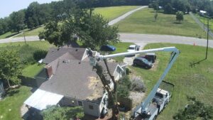 An aerial view of a worker in a bucket truck removing a tree near a house, with cut branches on the roof, by Crockett's Tree Service in Lafayette, IN.