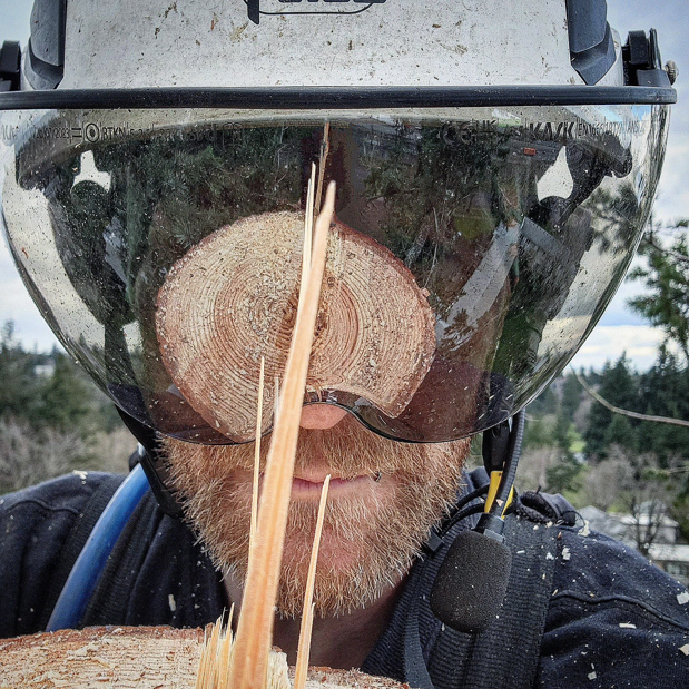 An arborist from Triple Acorn Tree Service in Portland, OR, with sawdust and a log reflection on their face shield after tree work.