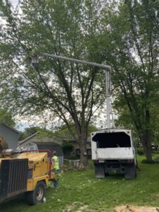 An arborist in a bucket truck actively pruning a large tree with a wood chipper nearby, performed by K.M.C Lawn & Tree service in Peoria, IL.