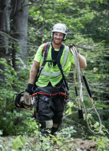 A smiling arborist in full safety gear, holding a chainsaw and ropes, in a wooded area for DJ's Tree Service in Colchester, VT.