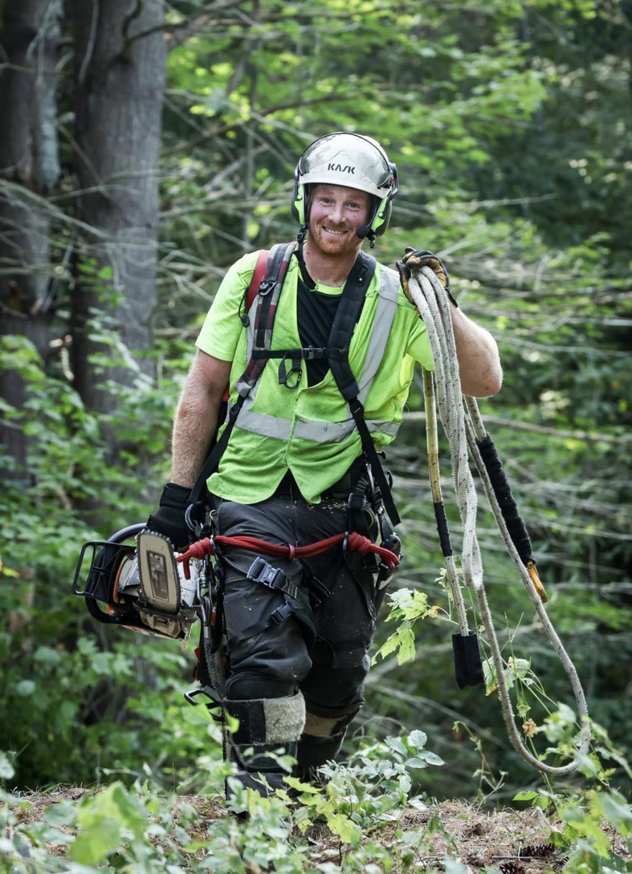 A smiling arborist in full safety gear, holding a chainsaw and ropes, in a wooded area for DJ's Tree Service in Colchester, VT.