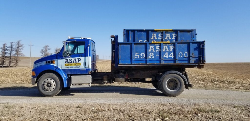 An ASAP Containers inc. truck with a blue dumpster on a dirt road, providing junk removal services in Omaha, NE.