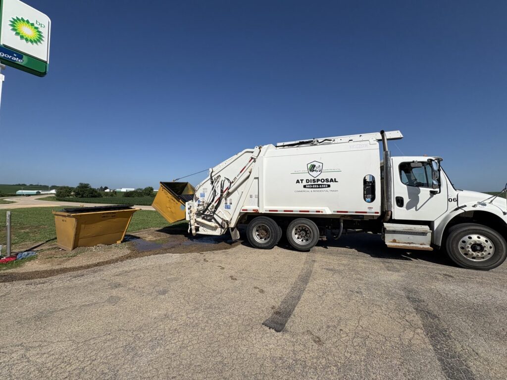 An AT Disposal garbage truck emptying a yellow commercial dumpster at a gas station in Dewitt, IA.
