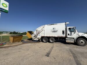 An AT Disposal garbage truck emptying a yellow commercial dumpster at a gas station in Dewitt, IA.