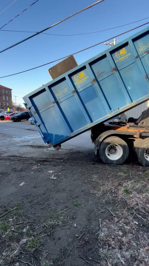 A blue AT Disposal roll-off dumpster being serviced, filled with junk including a mattress, in Dewitt, IA.
