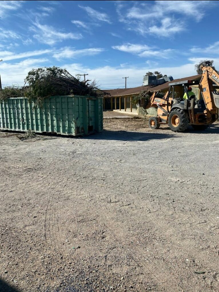 A backhoe loading tree branches and construction debris into a roll-off dumpster for A Track-Out Solution in Las Vegas, NV.