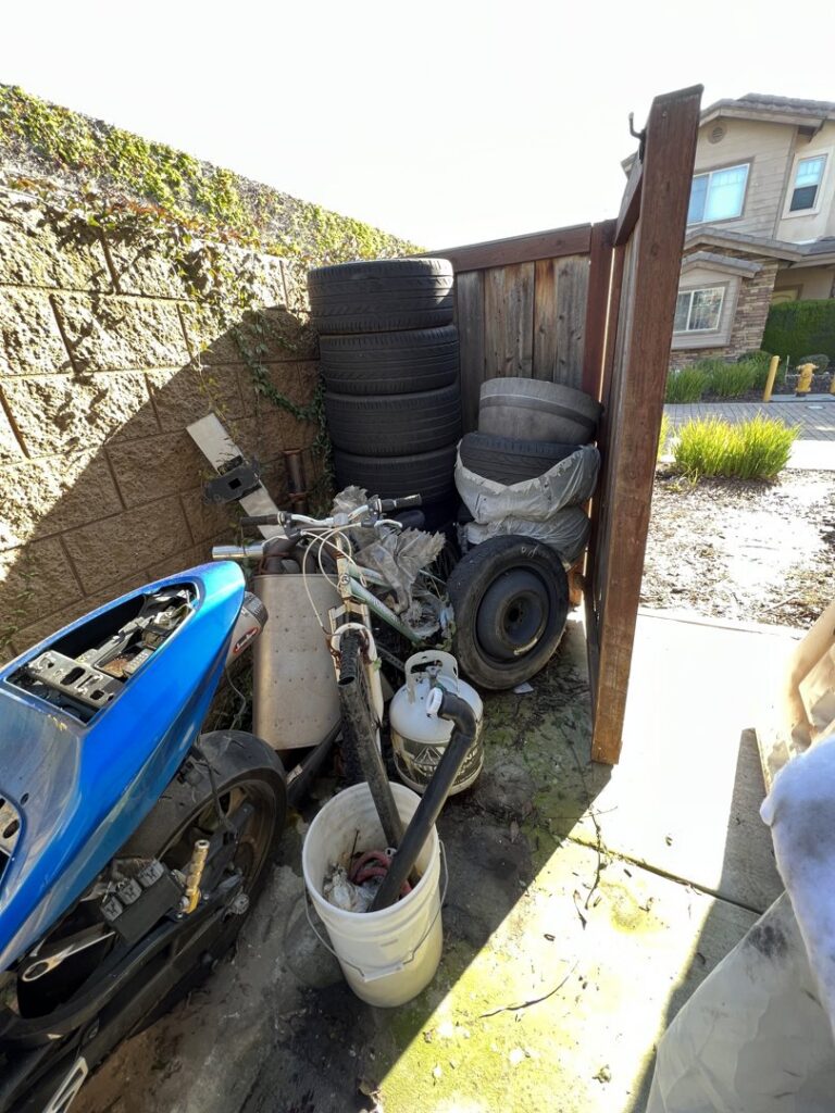 A backyard side area filled with tires, a bicycle, and other junk for removal by Impact Environmental Co. in El Cajon, CA.