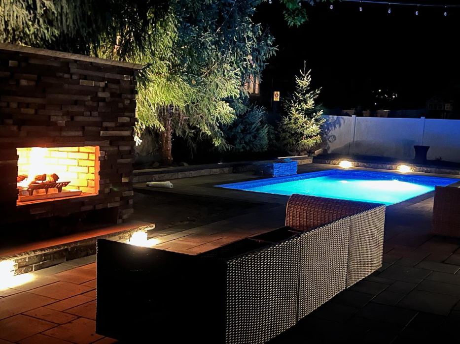 A stunning backyard oasis at night featuring a lit pool, outdoor fireplace, and patio by MVP Pools in Woodbridge, NJ