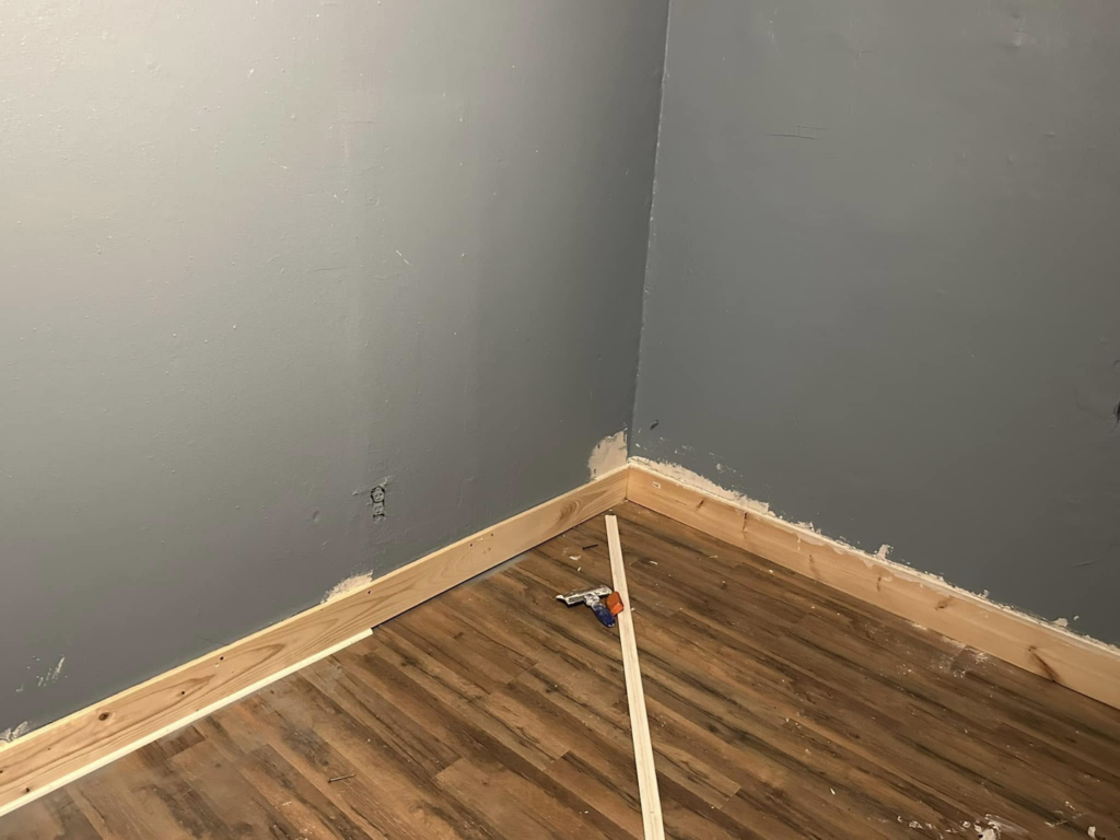 New baseboard trim being installed on a grey wall with wood-look flooring by Snell & Sons Construction LLC in Mobile, AL.