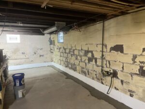 A basement wall showing peeling paint from water damage with a newly installed waterproofing barrier by TCM Waterproofing, LLC in Canton, OH.