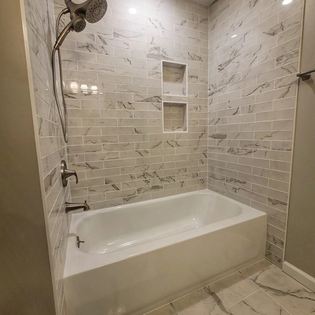 Bathroom with a bathtub and shower combo, featuring light grey marble-look subway tiles and two built-in niches by Supreme Tile & Flooring in Charlotte, NC.