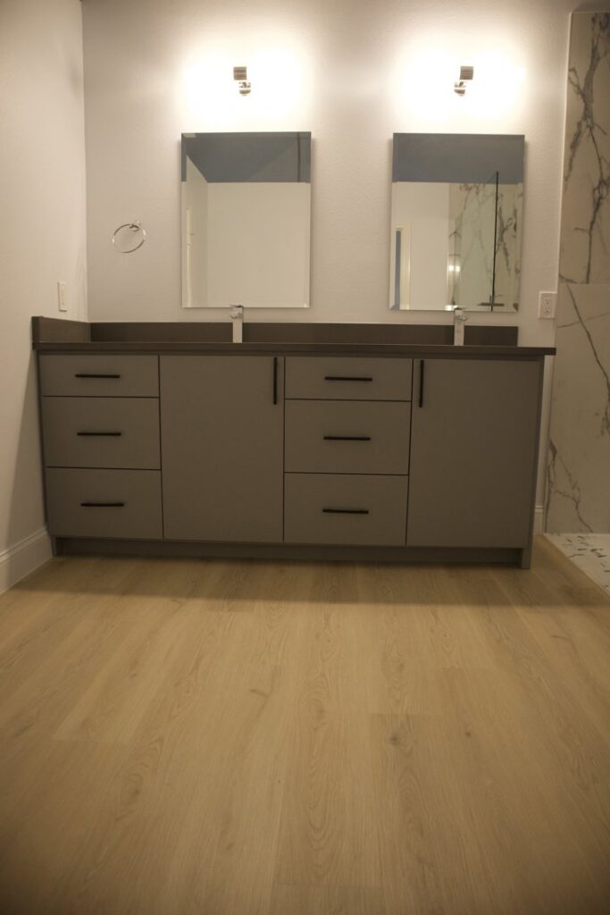 A renovated bathroom featuring a double vanity with grey cabinets and light wood-look flooring by Whole House Remodel & Design North Dallas Tx in Frisco, TX.