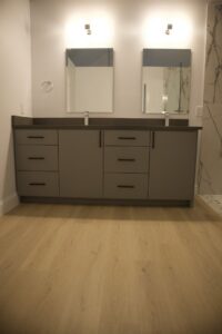 A renovated bathroom featuring a double vanity with grey cabinets and light wood-look flooring by Whole House Remodel & Design North Dallas Tx in Frisco, TX.