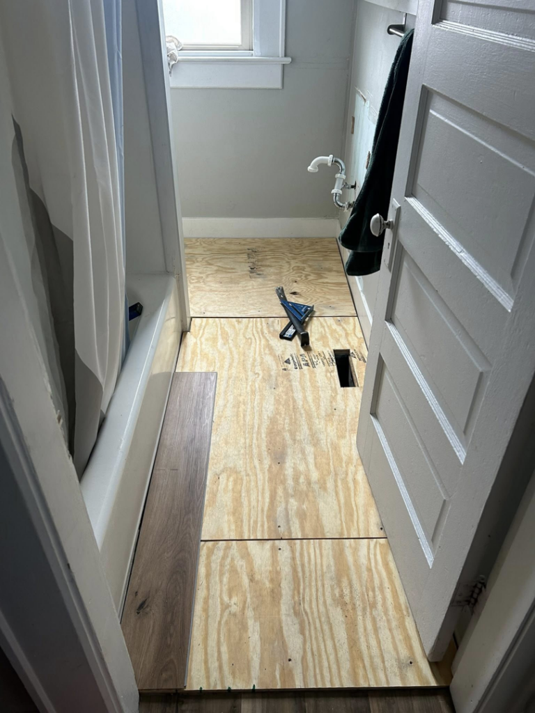 A bathroom showing flooring installation in progress with subfloor and a new plank, by CJ Flooring LLC in Flat Rock, MI.