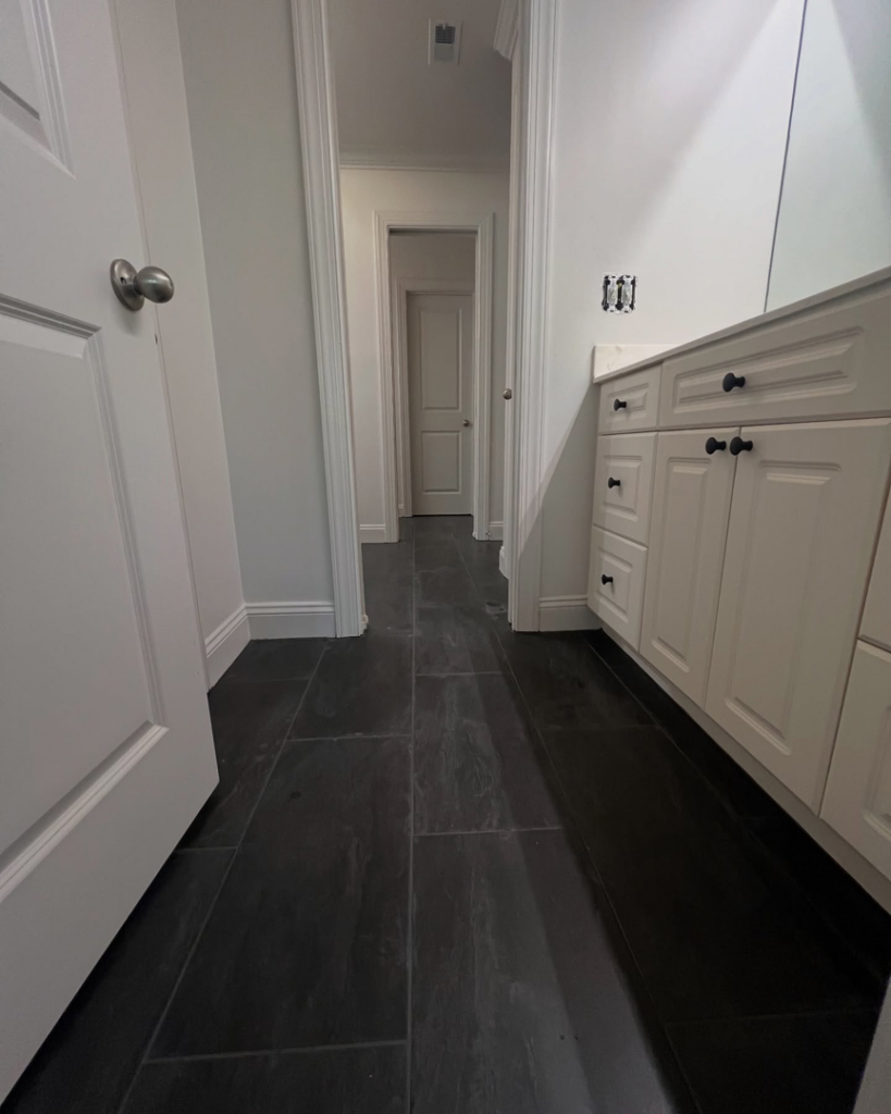 A renovated bathroom hallway with new dark floor tiles and a white vanity installation by TileWorks in Birmingham, AL.