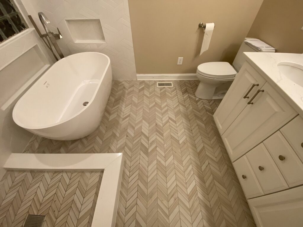 A renovated bathroom featuring herringbone pattern floor tiles and a white vanity by Goubar Tile And More in Raleigh, NC.