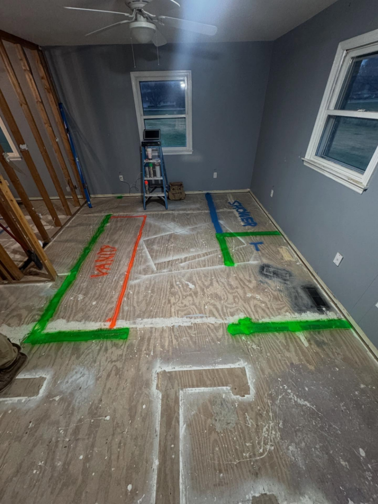 A room under renovation with 'VANITY' and 'SHOWER' marked on the plywood floor, showing bathroom layout by Shackelford Home Designs in Loveland, OH.
