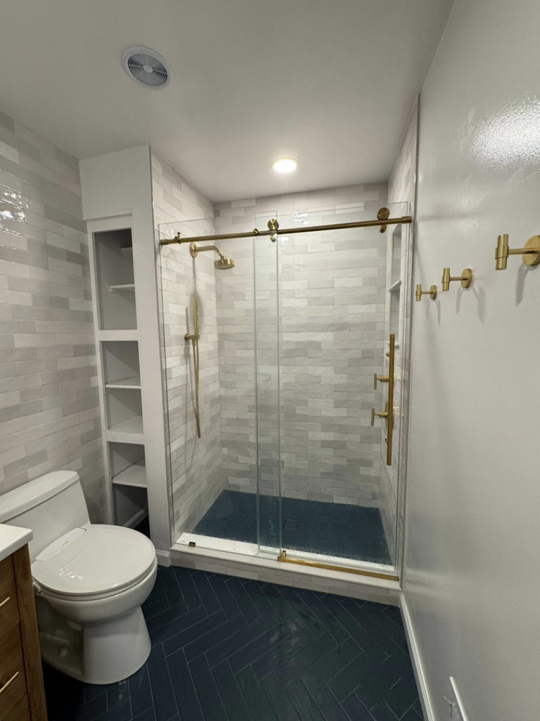A complete bathroom remodel featuring a toilet, built-in shelves, a shower with glass door, and herringbone floor tiles by NC Home Remodeling and Custom Cabinetry in Alexandria, VA.