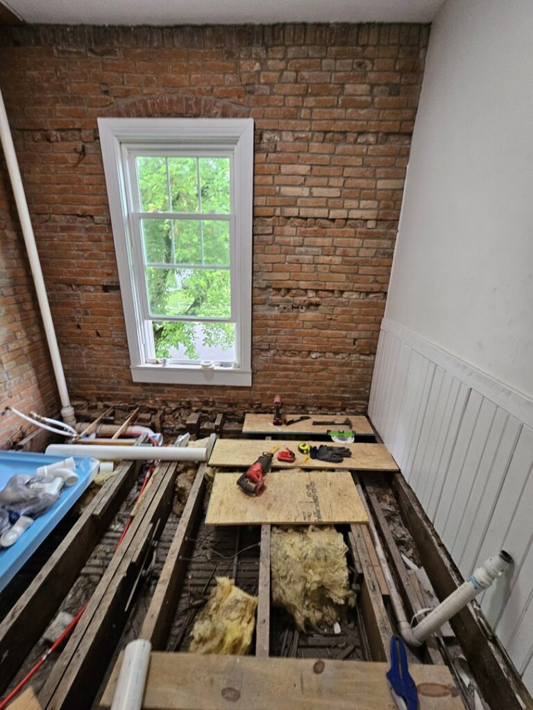 A bathroom renovation in progress showing exposed brick, subfloor, and plumbing work by Imperial Access Contracting in Evansville, IN.