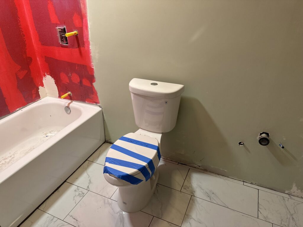 A bathroom under renovation, featuring a new toilet, tile flooring, and red waterproofing on the shower wall, by Jake and Jake Construction LLC in Sandy, UT.