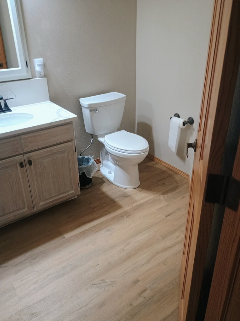 A newly renovated bathroom featuring a modern toilet, vanity, and wood-look flooring by The Cowboy Carpenter Handyman Service LLC in Bismarck, ND.