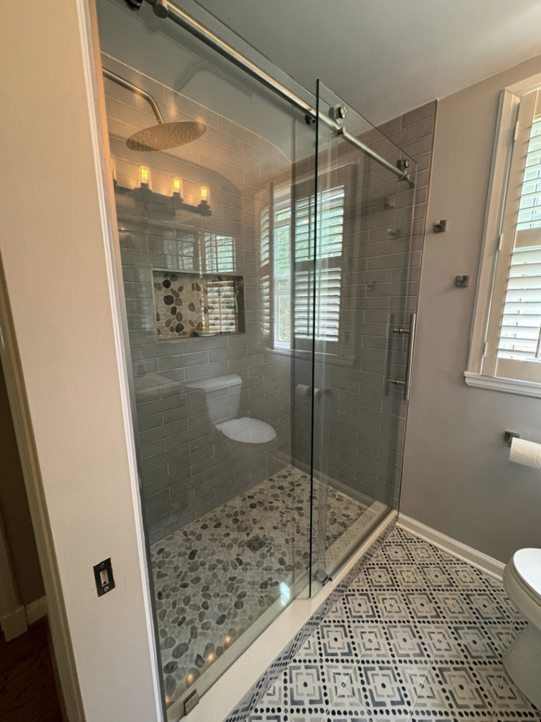 A modern bathroom shower with a glass sliding door, subway tiled walls, and a pebble floor, installed by NC Home Remodeling and Custom Cabinetry in Alexandria, VA.