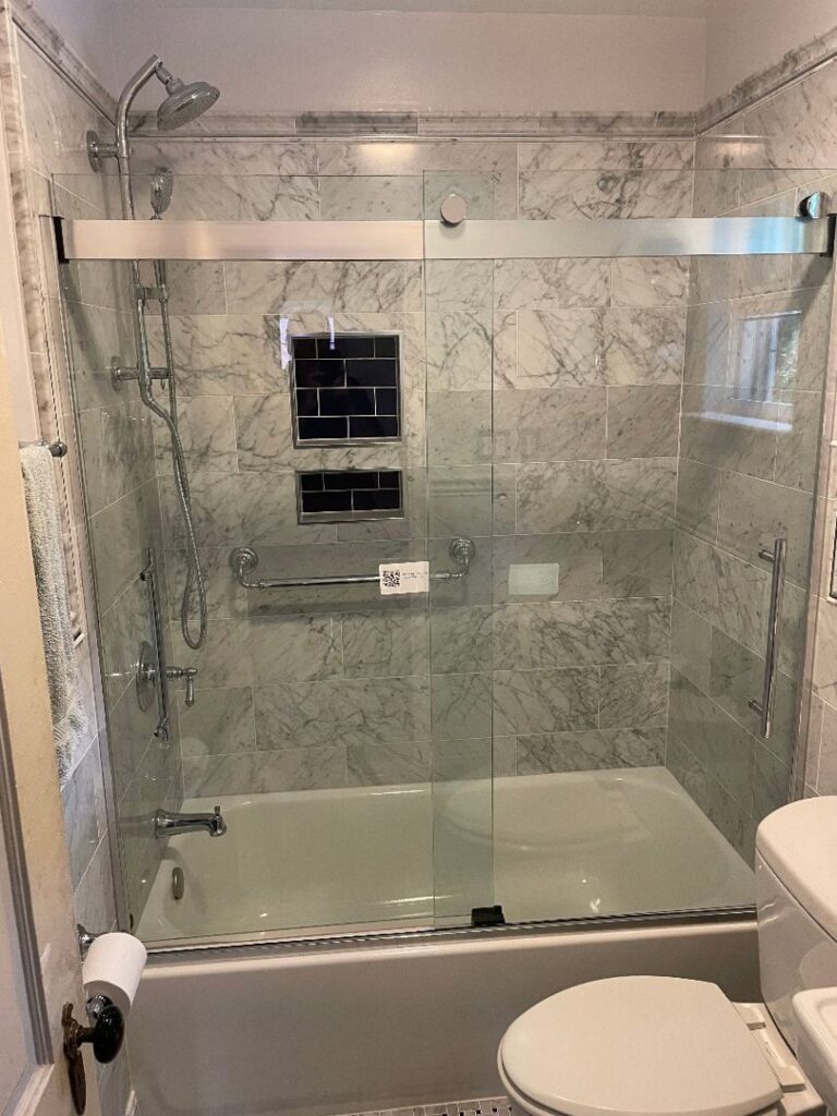 A renovated bathroom with a shower and tub combination, new marble-look tiles, and a modern glass shower door by Ruach Home Services in Laurel, MD.