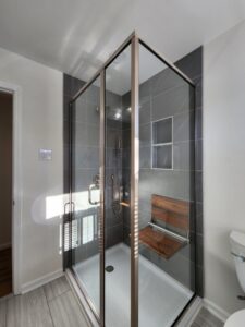Modern bathroom shower renovation with grey tiles, glass enclosure, and wooden seat by KC's Improvement & Construction Co., Inc. in Greensboro, NC