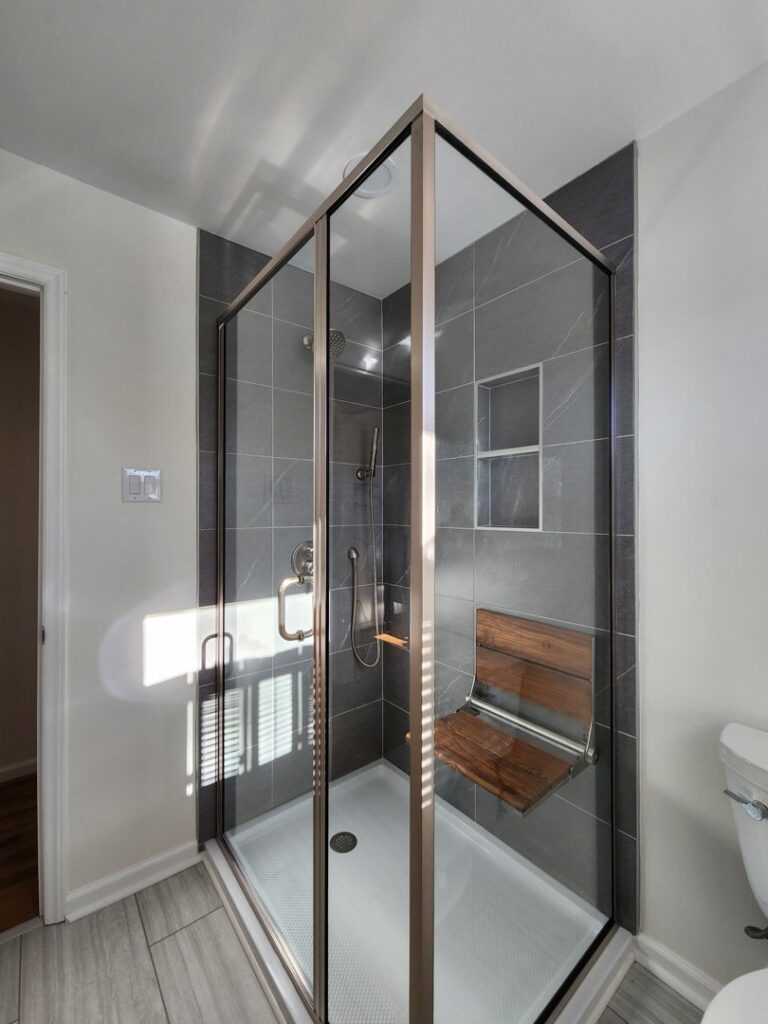 Modern bathroom shower renovation with grey tiles, glass enclosure, and wooden seat by KC's Improvement & Construction Co., Inc. in Greensboro, NC