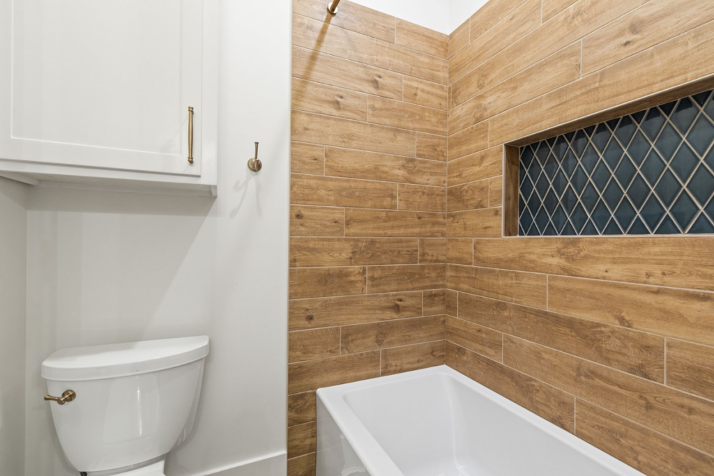 A bathroom featuring a new toilet, bathtub, wood-look wall tiles, and a tiled niche installed by Dugas Construction, LLC in Lafayette, LA.