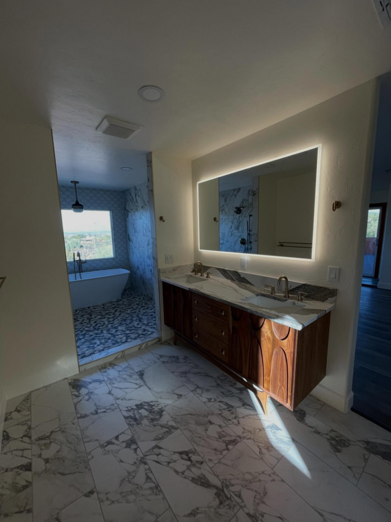 A renovated bathroom with a double vanity, illuminated mirror, and marble-look floor tiles by Raven Construction LLC in Tucson, AZ.
