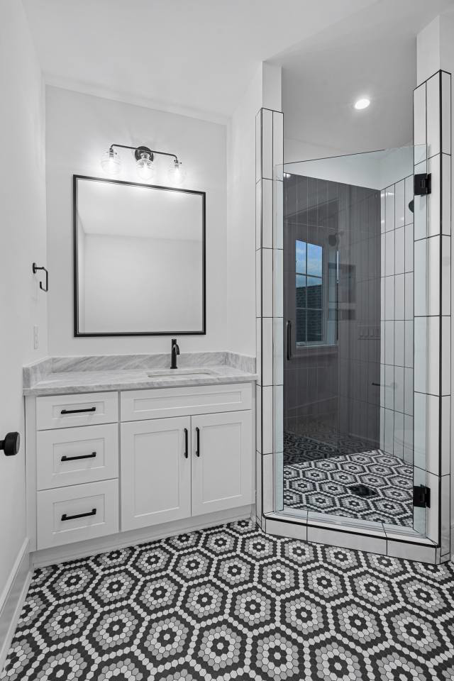 A modern bathroom featuring a white vanity, black mirror, and walk-in shower with hexagonal floor tiles by CML Builders in Charlotte, NC.