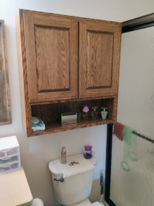 A wooden wall cabinet installed above a toilet in a bathroom by The Cowboy Carpenter Handyman Service LLC in Bismarck, ND.