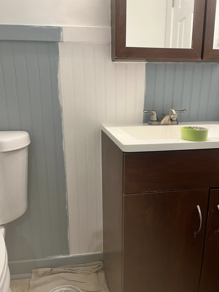 A bathroom wall with beadboard paneling being painted light blue/grey, showing work in progress by Coach Rose Painting Team in Norfolk, VA.