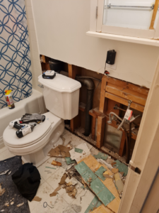 A bathroom wall opened up for plumbing repair, showing exposed pipes and studs, with tools on the toilet, by All American Handyman services in Billings, MT