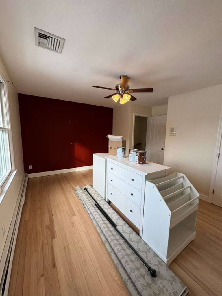 A bedroom featuring a newly painted red accent wall and paint cans, showcasing an interior painting job by Quinonez Painting & Renovations LLC in Providence, RI.