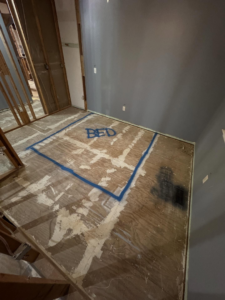 A room under renovation with 'BED' marked on the plywood floor, indicating bedroom layout planning by Shackelford Home Designs in Loveland, OH.