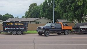 A BeeBop's Junk Removal truck towing two trailers, one branded for junk removal, parked on a street in Sioux Falls, SD.