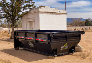 A black dump trailer from Duke City Dumpster Rentals LLC parked in a dirt lot next to a mausoleum, indicating a cleanup or junk removal job in Albuquerque, NM.