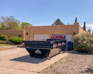 A black dump trailer from Duke City Dumpster Rentals LLC in a residential driveway with a mattress leaning against the garage, ready for junk removal in Albuquerque, NM.
