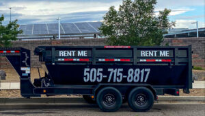 A black dump trailer with 'RENT ME' branding from Duke City Dumpster Rentals LLC, available for rent in Albuquerque, NM, with solar panels in the background.