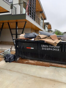 A black dumpster overflowing with cardboard and debris from Liberty Hill Dumpster Rental at a modern building in Austin, TX