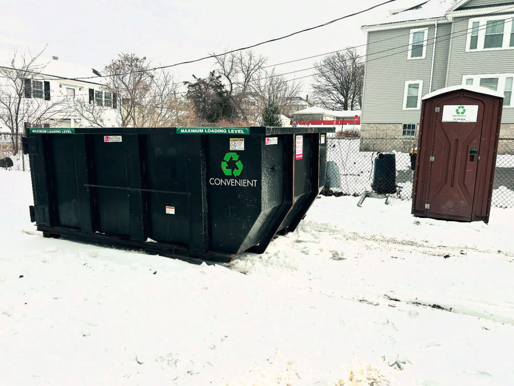 A black dumpster and a portable toilet placed in a snowy residential yard for disposal services from Convenient Disposal & Sanitation in Fall River, MA.