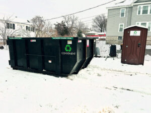 A black dumpster and a portable toilet placed in a snowy residential yard for disposal services from Convenient Disposal & Sanitation in Fall River, MA.