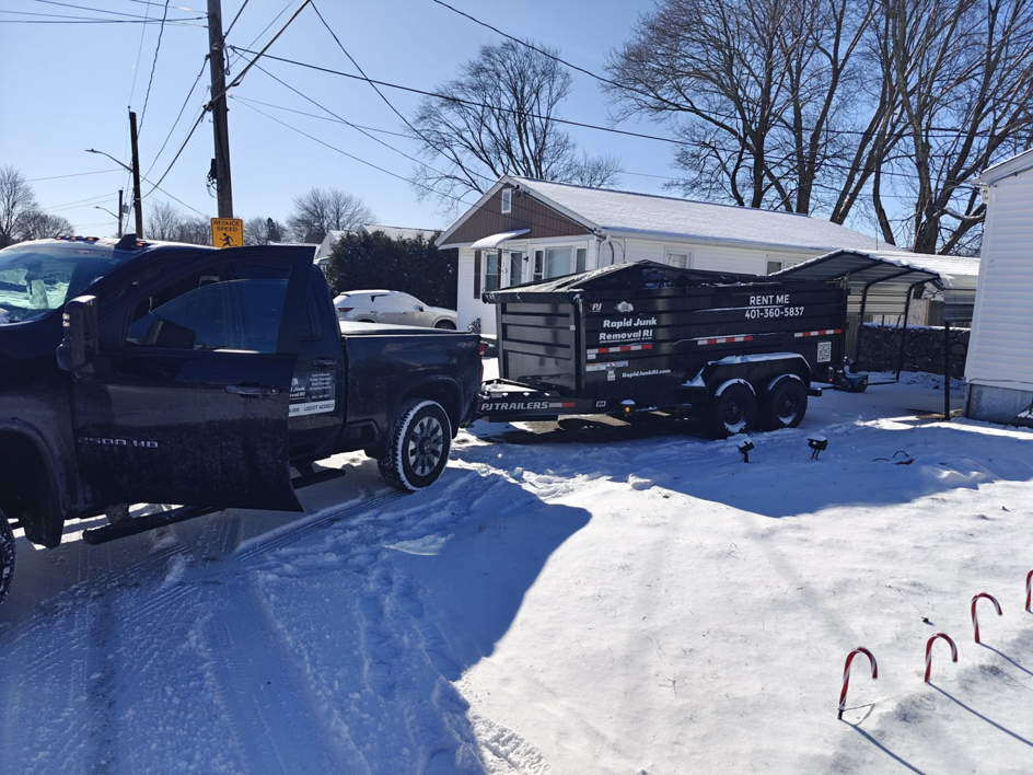 A black pickup truck towing a dumpster trailer from Dumpsters RI.com - Dumpster Rentals RI in a snowy residential area in Rhode Island.