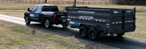 A black pickup truck towing an empty Mobiledumps dumpster trailer on a road for Mobiledumps Northwest Arkansas in Rogers, AR.