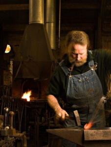 A skilled blacksmith forging hot metal on an anvil at Bighorn Forge, Inc. in Kewaskum, WI, demonstrating custom metalwork capabilities.