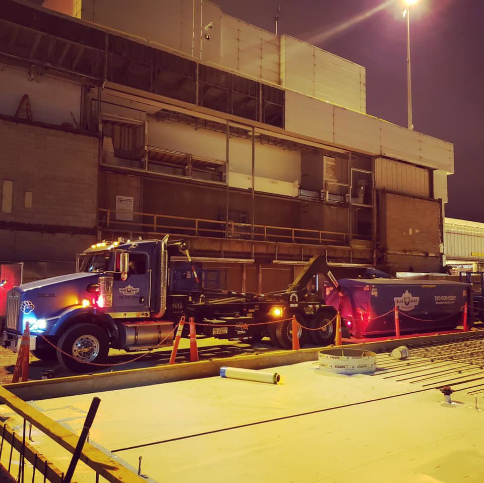 A Blue Bear Waste Services truck with a dumpster at a construction site at night in Denver, CO.